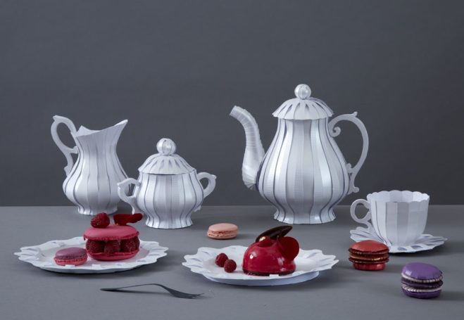 Elegantes weißes Teeservice aus Papier mit Kuchen und Macarons auf einem grauen Tisch.